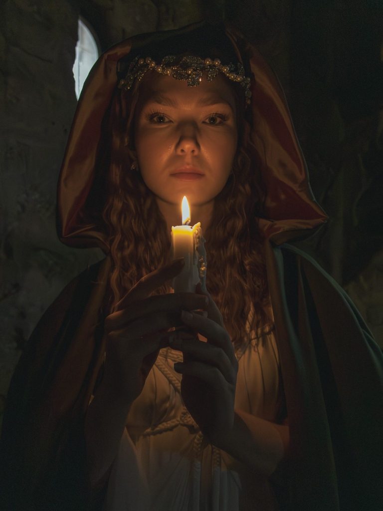 Junge Frau in historischem Gewand hält eine Kerze in dunkler Umgebung.